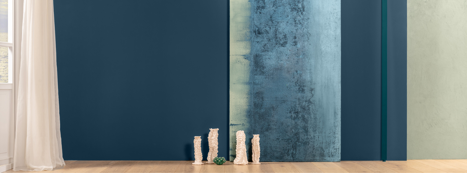 Ein moderner Innenraum mit drei Wänden in verschiedenen Farbtönen: eine dunkle blaue Wand, eine vertikale Wand mit einem sanften, strukturierten Verlauf in Blau und eine grüne Wand. Vor den Wänden stehen mehrere dekorative Objekte in Form von schlanken, weißen Vasen und einer kleinen Grünpflanze auf einem hellen Holzboden. 